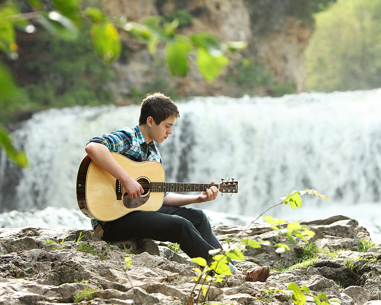 Young man playing a guitar in front of a waterfall in Wisconsin for his senior portraits with Kari Layland Photography